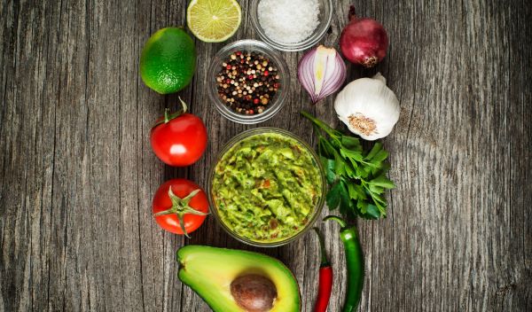 een kommetje met guacamole op tafel en ingredienten om het te maekn